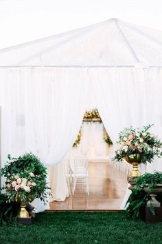 an outdoor wedding setup with white drapes and greenery on the grass, surrounded by potted plants