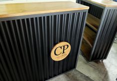 two wooden tables with black corrugated coverings and a logo on the front one has a circular wood plaque that reads capital