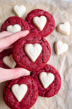 red velvet cookies with white heart shaped frosting on parchment paper next to two hands