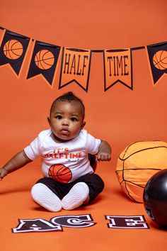 a baby sitting on the floor in front of basketball balls and bunting banner with name time