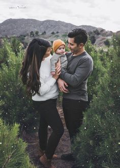 a man and woman holding a baby in their arms while walking through some trees with mountains in the background