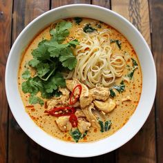 a white bowl filled with noodles and chicken on top of a wooden table next to cilantro