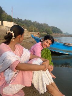 two women are sitting on the shore with boats in the background