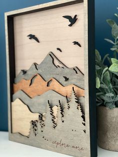 a wooden frame with birds flying over mountains and trees in the background, on a table next to a potted plant