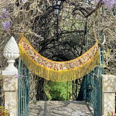 an iron gate with a yellow fringe hanging from it's sides, surrounded by flowers and trees