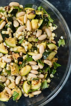 a glass bowl filled with beans and greens on top of a blue countertop next to a wooden spoon