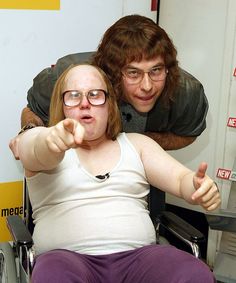 a woman in a wheel chair pointing at the camera with a man standing behind her