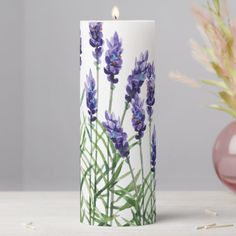 a white candle with purple flowers on it next to a pink vase and flower arrangement