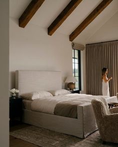 a woman in a white dress is standing near a large bed and chair, looking out the window