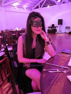 a woman sitting at a table wearing a masquerade and holding her hand to her face