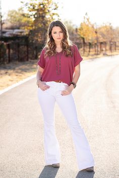 a woman standing on the side of a road wearing white pants and a maroon shirt