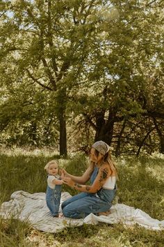 a woman sitting on top of a blanket holding a baby in her lap next to trees