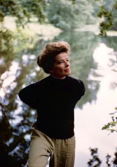 a woman standing in front of a body of water with trees around her and looking off into the distance