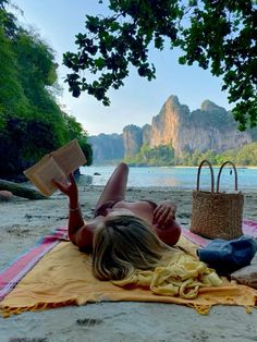 a woman laying on top of a beach next to a basket and book in her hand