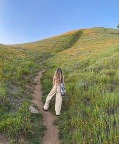 a woman walking down a dirt path in a field with wildflowers on the hillside