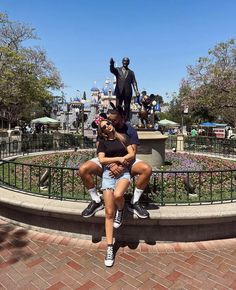 a man and woman are sitting on a bench in front of the statue of mickey mouse