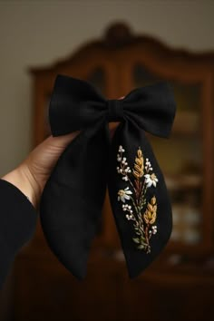 a hand holding a black bow with flowers and leaves embroidered on the side, in front of a mirror