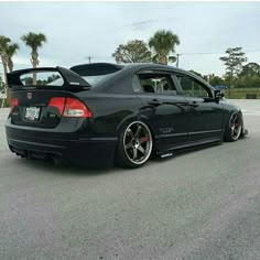 the rear end of a black car parked in a parking lot next to palm trees