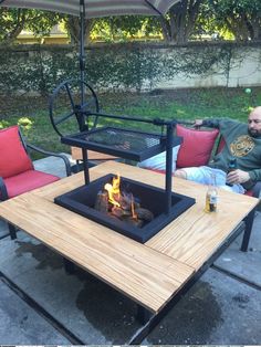 a man sitting in front of a fire pit on top of a wooden table with an umbrella over it