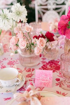 pink and white flowers are in vases on a table with plates, cups and saucers