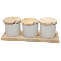 three white ceramic containers with wooden lids and spoons on a wood tray against a white background