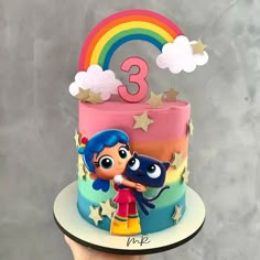 a hand holding up a cake with the number three on it and two cartoon characters