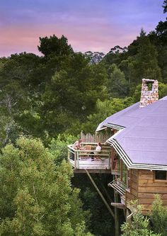 two people sitting on the porch of a wooden cabin in the woods with trees surrounding them