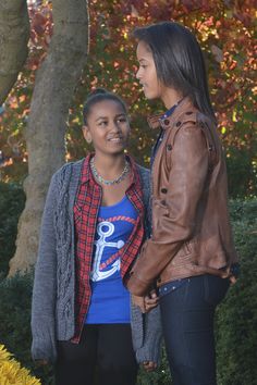 two young women standing next to each other in front of trees and bushes with orange leaves