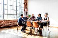 four people sitting at a table in an office setting, with one person standing up