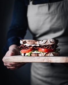 a person in an apron holding a wooden tray with a sandwich on top of it