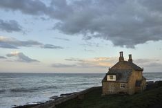 an old house sitting on the edge of a cliff by the ocean with dark clouds in the sky