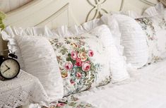 a white bed topped with lots of pillows next to a night stand and alarm clock