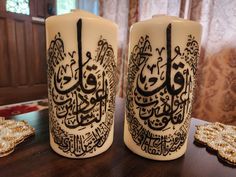 two white candles decorated with arabic calligraphy on top of a table next to cookies