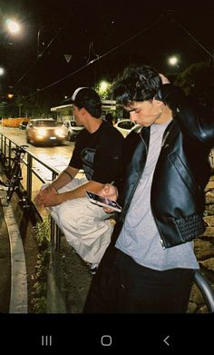 two young men standing next to each other on a sidewalk at night with their cell phones in their hands