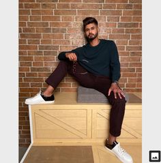 a man sitting on top of a wooden dresser next to a brick wall with his hands in his pockets