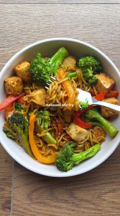 a white bowl filled with noodles, broccoli and tofu stir fry on top of a wooden table