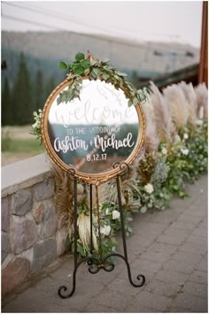 a welcome sign with greenery on it is sitting next to a stone wall and flowers