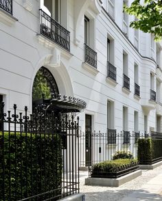 a white building with black iron fence and bushes on the sidewalk in front of it