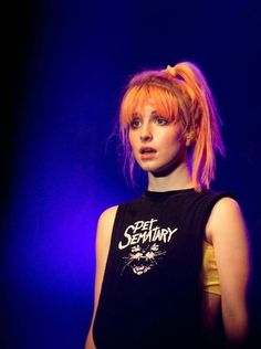 a woman with orange hair and bangs standing in front of a blue background wearing a black tank top