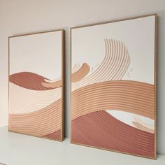 two framed art pieces on a shelf in front of a white wall with pink and beige waves