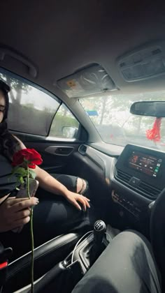 a woman sitting in the driver's seat of a car holding a red rose