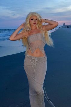 a woman standing on top of a sandy beach next to the ocean at sunset with her hands behind her head