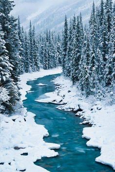 a river surrounded by snow covered trees in the middle of a snowy mountain area with blue water