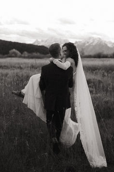 a bride and groom walking through the grass in an open field with mountains in the background