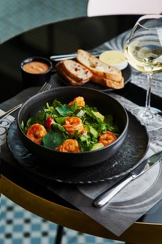 a bowl of shrimp and greens on a table with bread, wine glasses and utensils