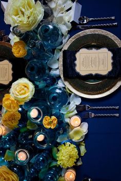 a table topped with plates and candles next to a flower arrangement on top of a blue table cloth