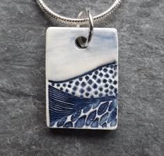 a blue and white tile pendant on a silver chain with an ocean scene in the background