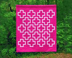 a pink quilt with white squares on it in front of some green trees and bushes