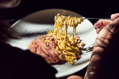 a person is eating some noodles on a white plate with a fork and knife in their hand