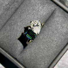 an engagement ring with green and blue stones in a velvet box on top of a table